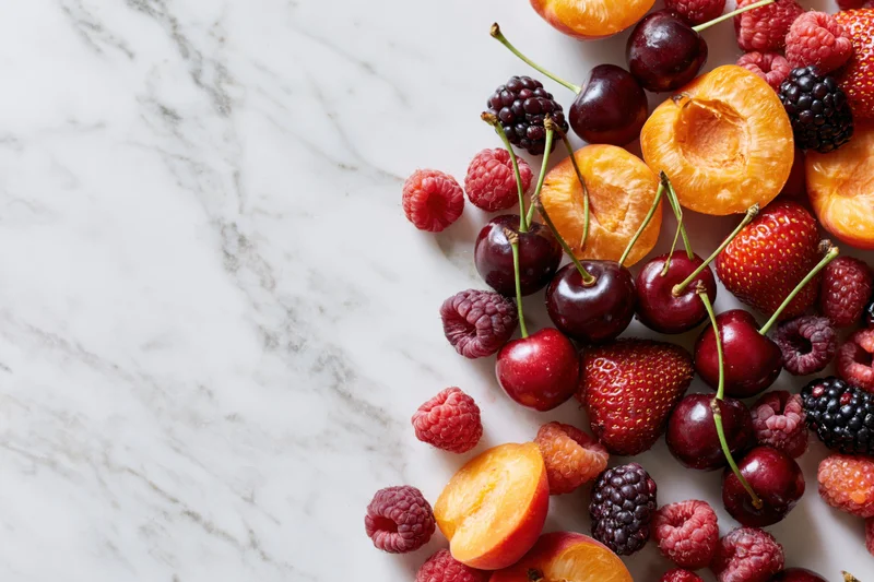 Halved apricots, cherries, strawberries, raspberries and blackberries arranged on a marble countertop with copyspace on the left