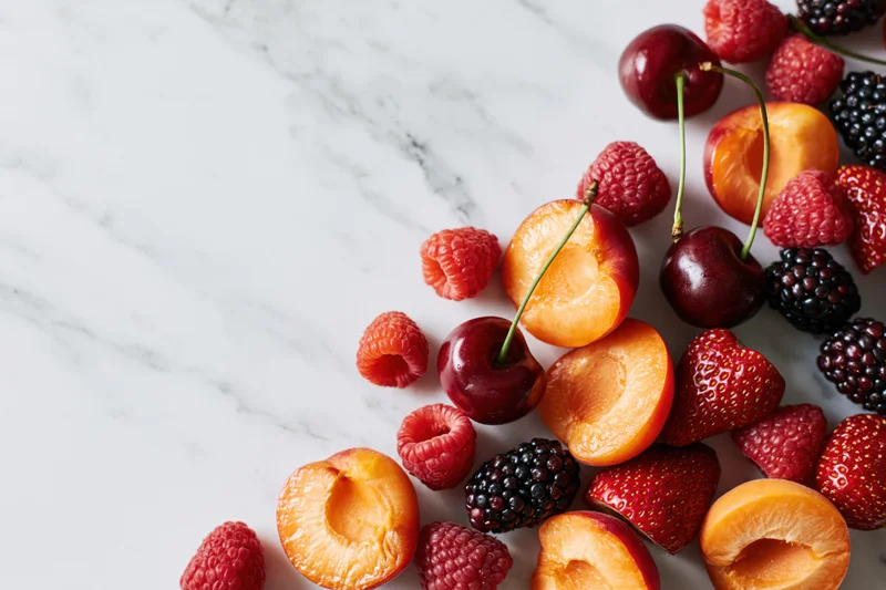 Halved apricots, cherries, strawberries, raspberries and blackberries arranged on a white marble surface