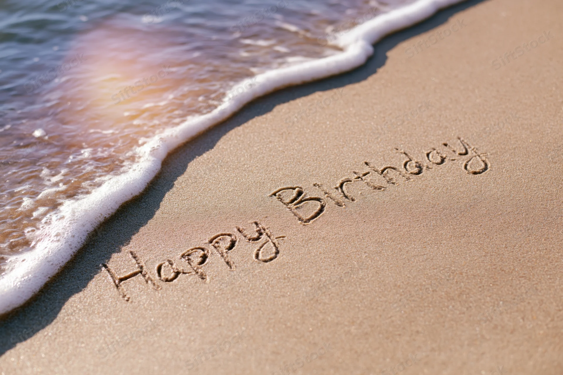 Happy birthday written in cursive on wet sand at beach shoreline with wave approaching