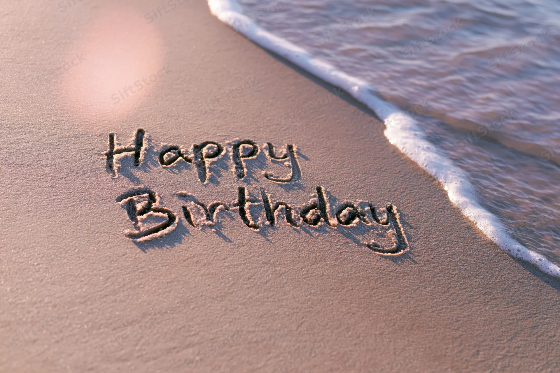 Happy birthday written in cursive on wet sand at beach with wave approaching