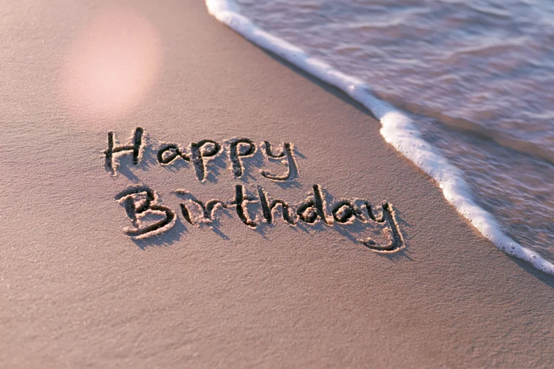 Happy birthday written in cursive on wet sand at beach with wave approaching
