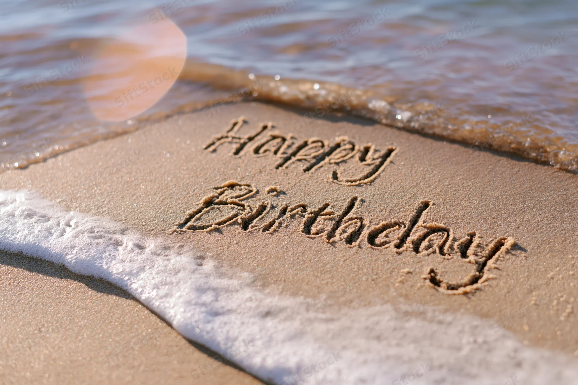 Happy birthday message written in cursive script on wet sand at beach shoreline with waves approaching