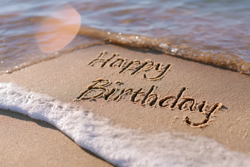 Happy birthday message written in cursive script on wet sand at beach shoreline with waves approaching