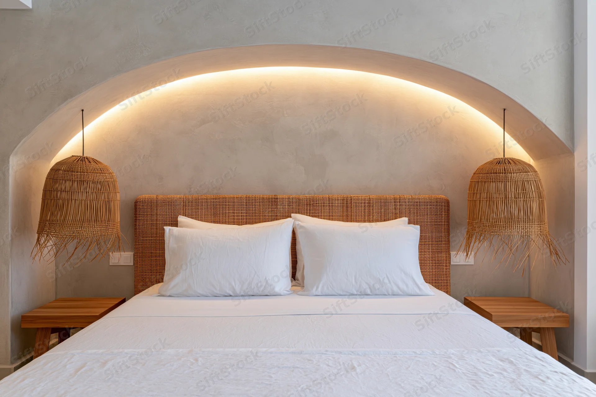 Double bed with white bedding and wicker headboard in a hotel room with arched recessed lighting and pendant lamps