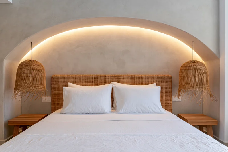 Double bed with white bedding and wicker headboard in a hotel room with arched recessed lighting and pendant lamps
