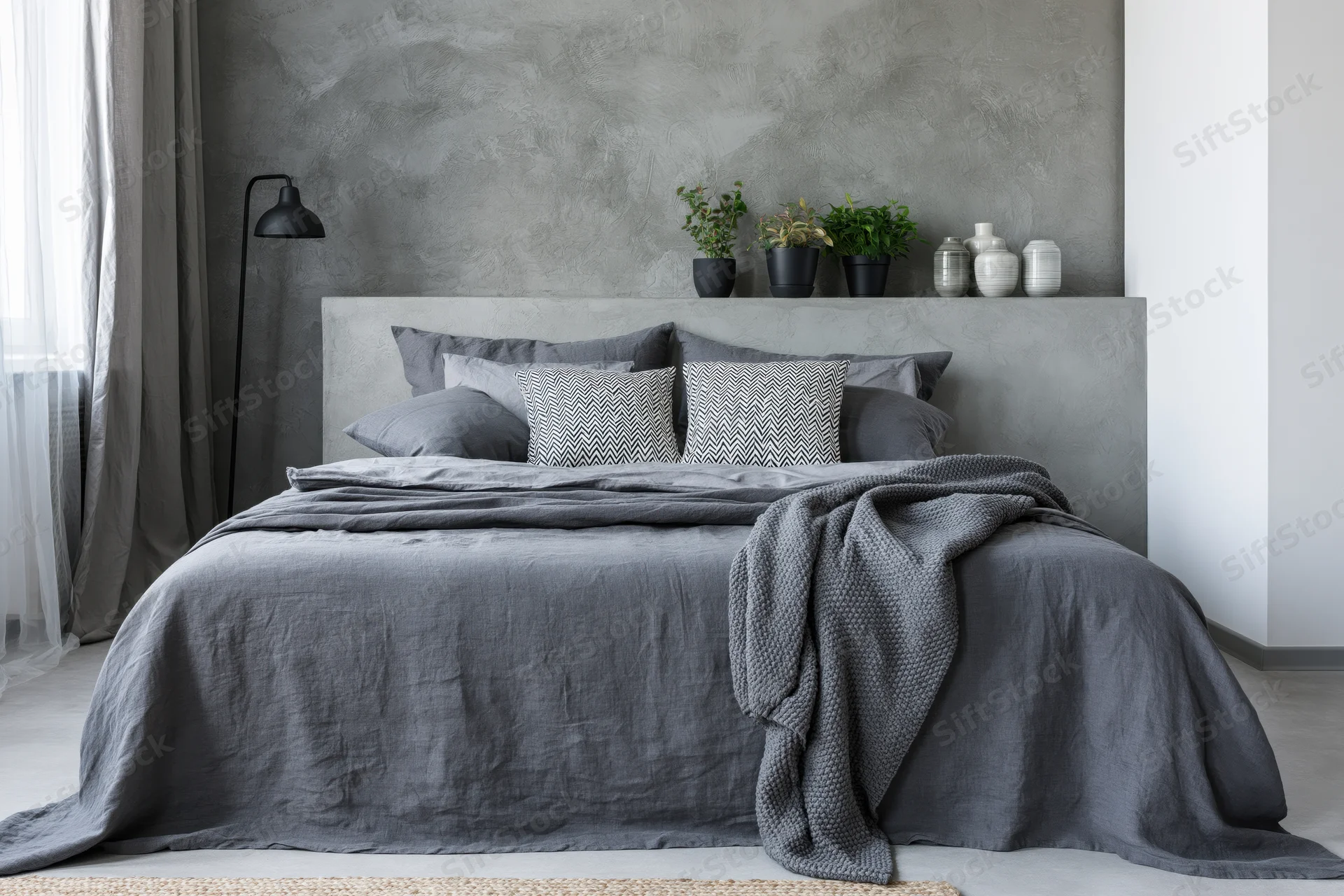 Industrial-style bedroom with gray concrete headboard and bedding, featuring plants and a floor lamp near a window