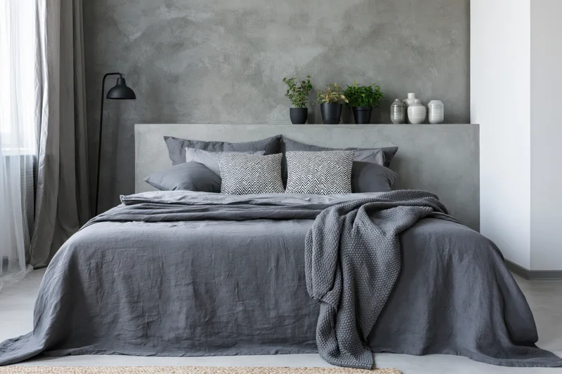 Industrial-style bedroom with gray concrete headboard and bedding, featuring plants and a floor lamp near a window