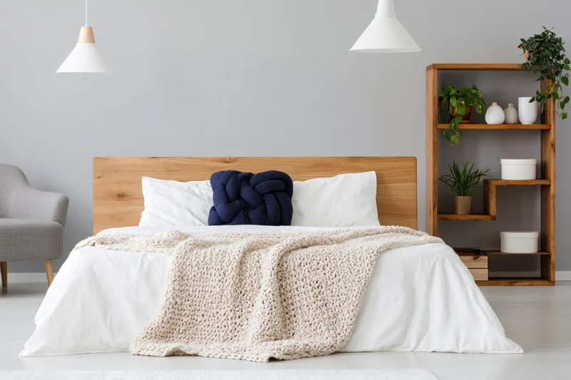 Minimalist bedroom with wooden bed and white bedding, featuring knit throw and shelving unit with plants