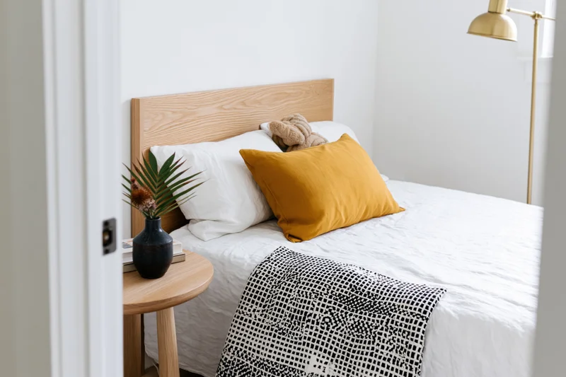 Minimalist bedroom interior viewed from doorway showing single bed with white bedding, mustard pillow, wooden headboard, and side table with vase and plant