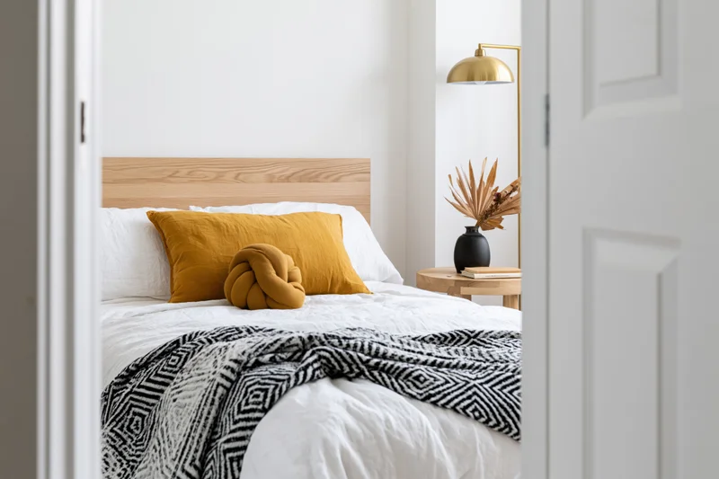 View through doorway into minimalist bedroom with single bed, white bedding, mustard pillow, and gold lamp beside wooden side table