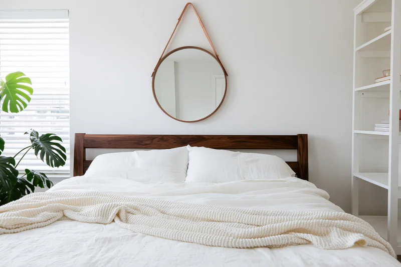Minimalist bedroom with dark walnut bed frame, white bedding, round mirror, and monstera plant near window