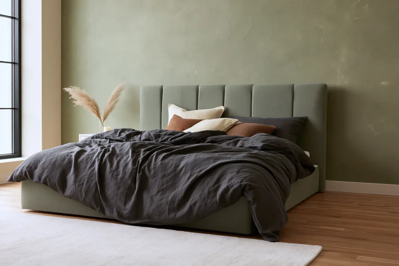 Low platform bed with dark bedding and green upholstered headboard in minimalist bedroom with wooden floor and window