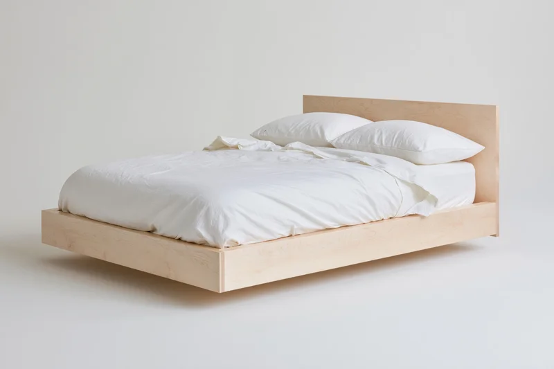 Minimalist platform bed with pale birch wood frame and white bedding on plain background