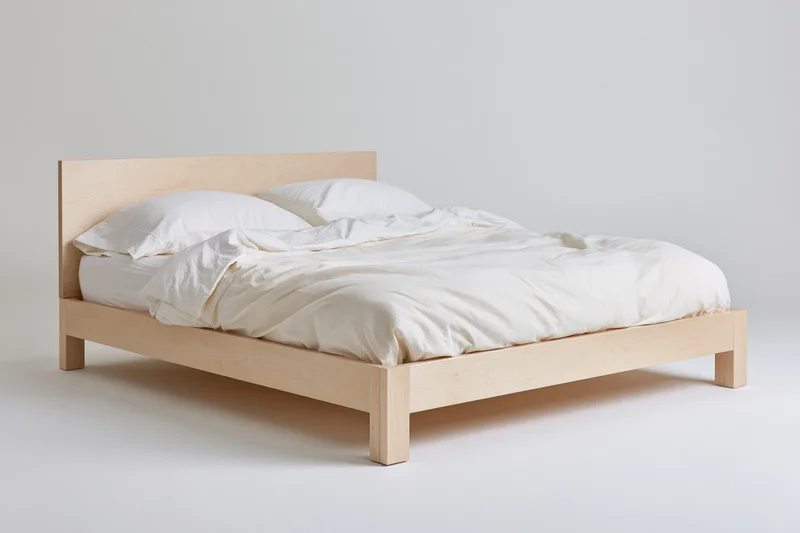 Minimalist platform bed with pale birch wood frame and white bedding on plain white background