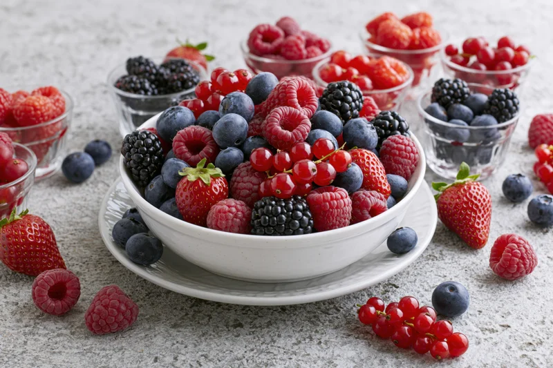 A white bowl filled with mixed fresh berries on a plate with scattered fruit around on a textured surface