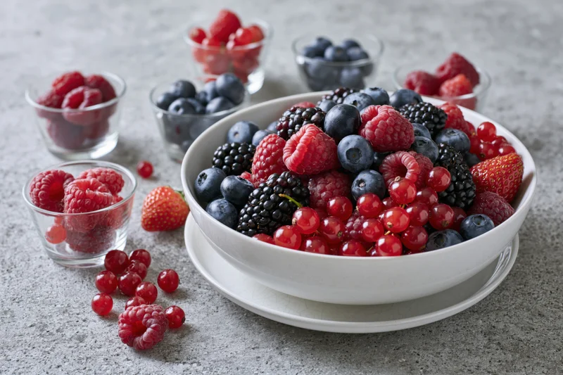 A white bowl filled with mixed fresh berries including raspberries, blueberries, blackberries and strawberries on a gray surface with small glass bowls and scattered fruit around