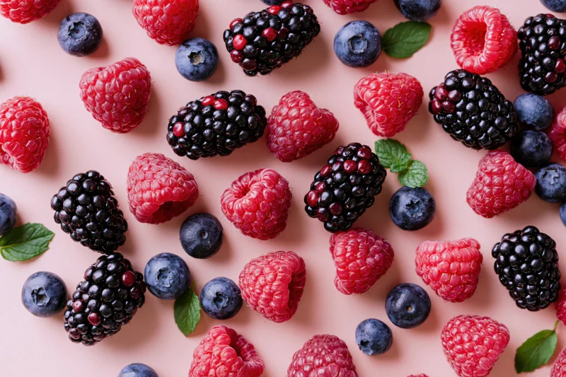 Mixed fresh berries including raspberries blackberries and blueberries arranged on a pink surface with mint leaves in a flat lay top view