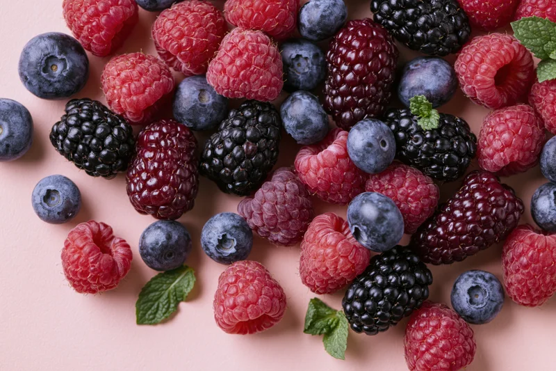 Mixed fresh raspberries blueberries and blackberries arranged on a pink surface with mint leaves