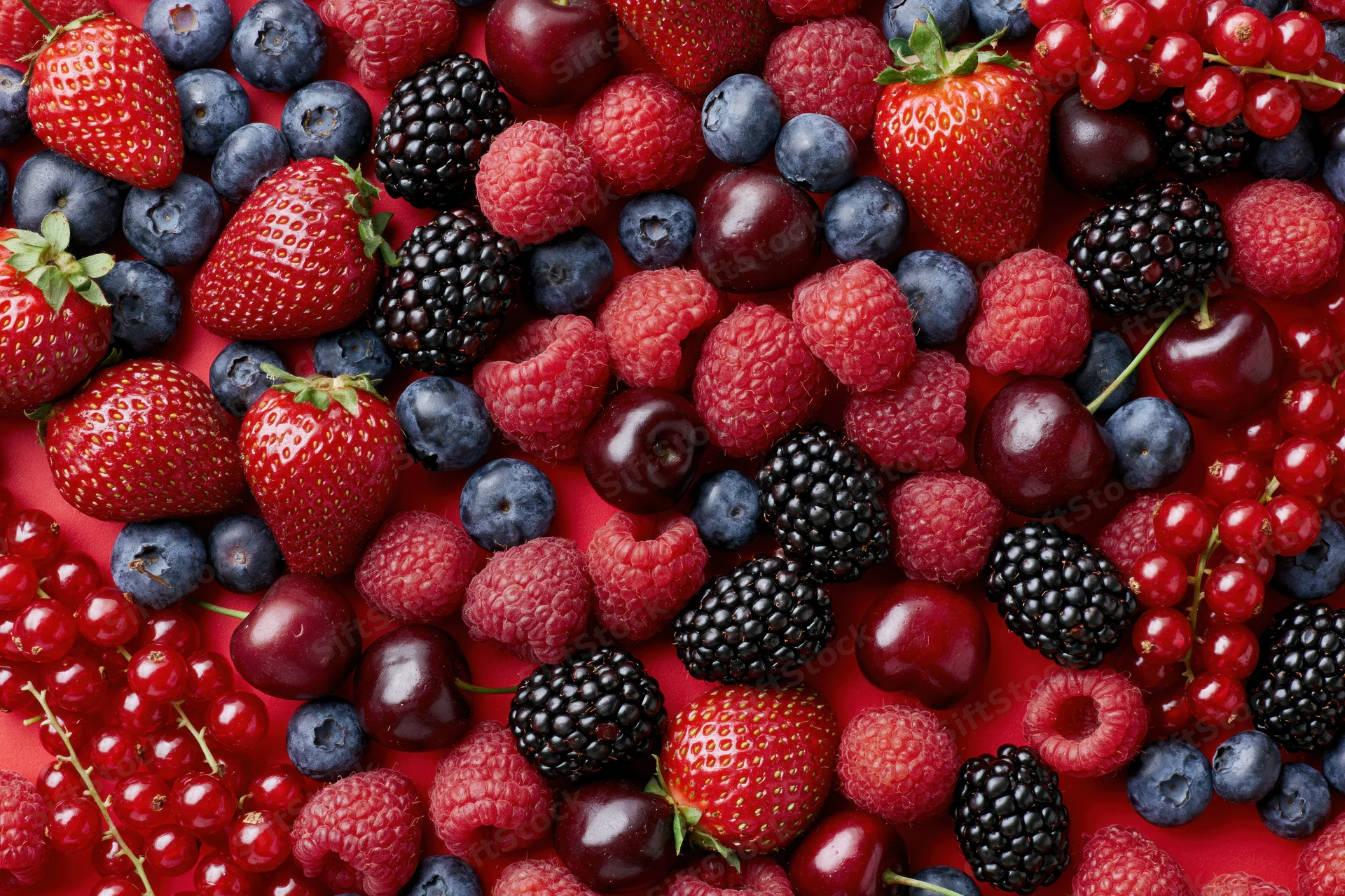 Mixed fresh berries scattered densely on a bright red surface in a flat lay top view