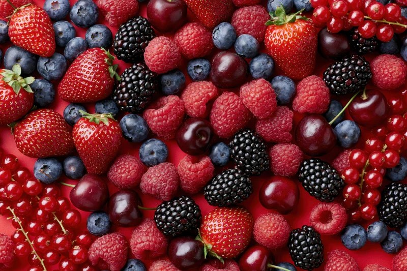 Mixed fresh berries scattered densely on a bright red surface in a flat lay top view