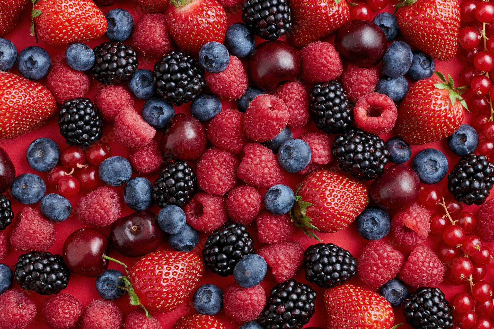 Mixed fresh berries scattered densely on a bright red surface in a flat lay top view