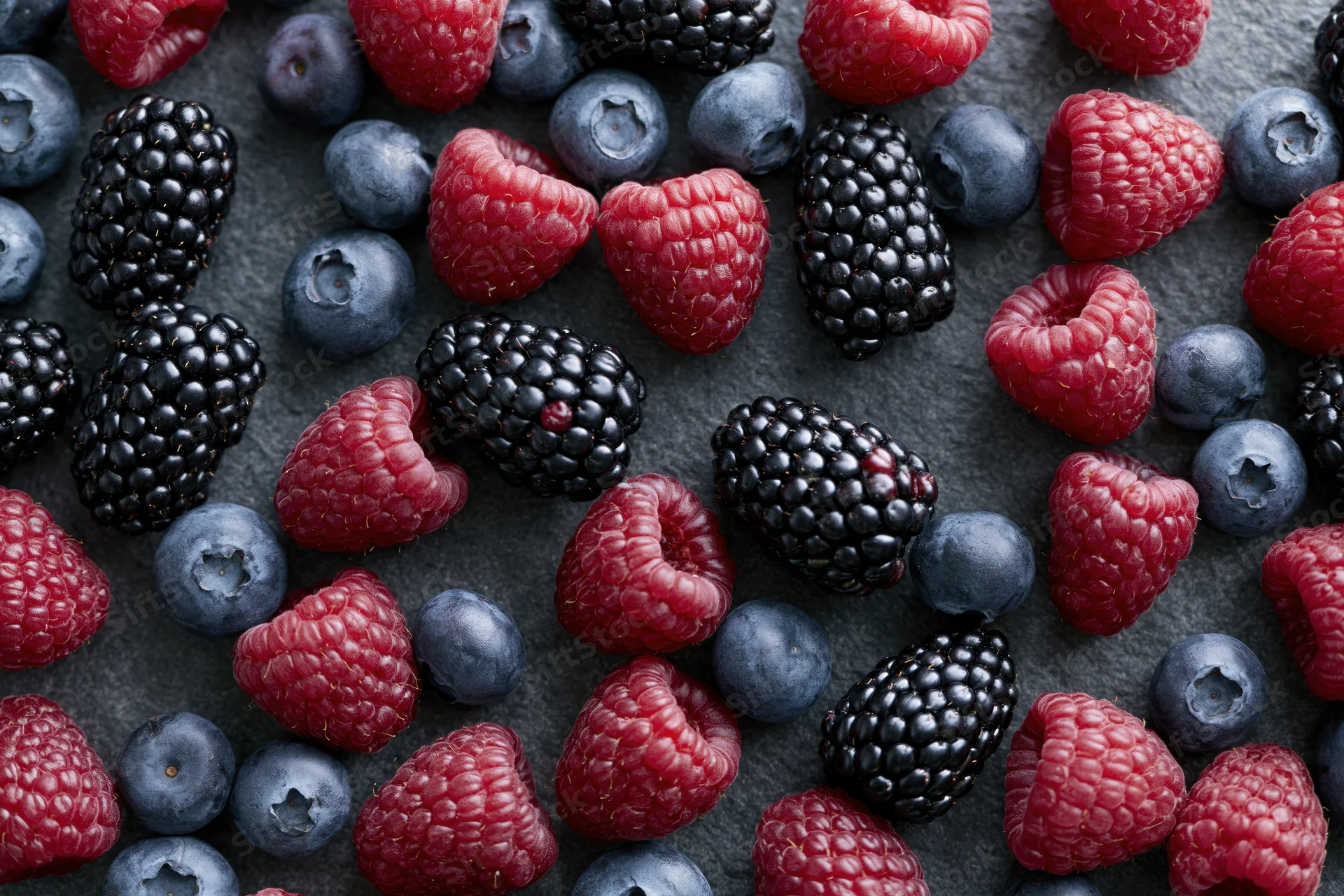 Mixed fresh berries scattered loosely across dark matte stone surface
