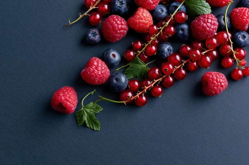 Fresh raspberries blueberries and redcurrants scattered on dark charcoal surface with leaves and stems