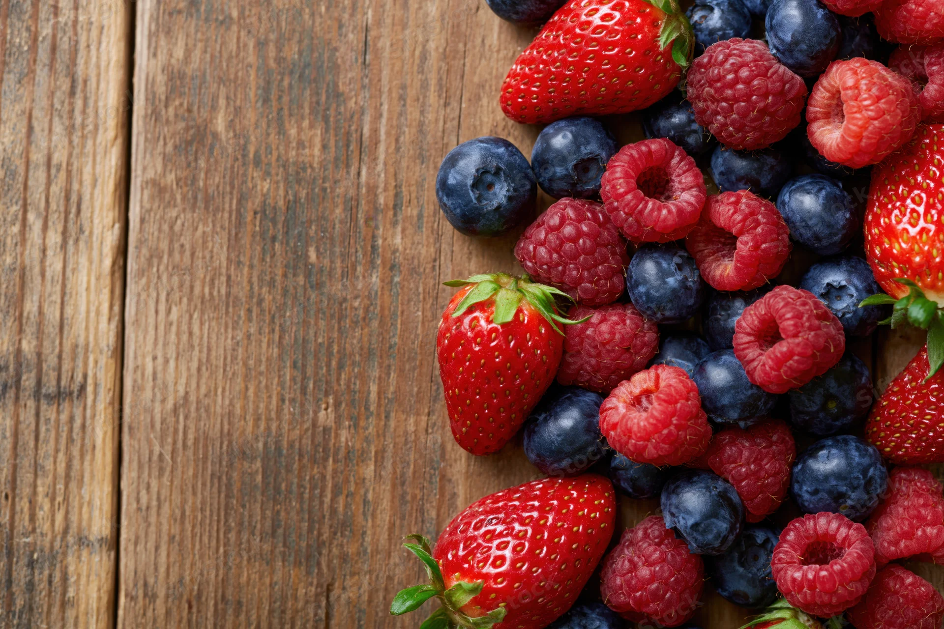 Mixed fresh strawberries blueberries and raspberries arranged on a wooden surface with copyspace on the left