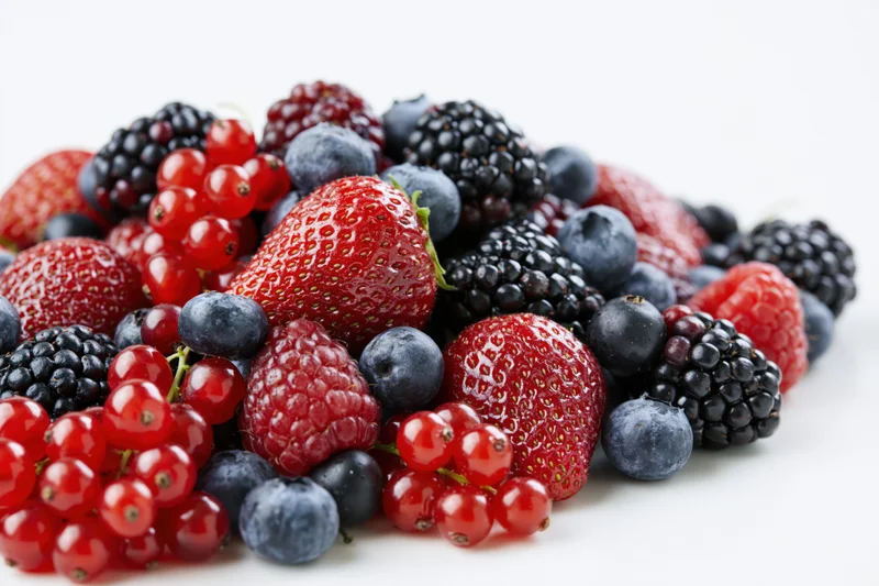 A pile of mixed fresh berries including strawberries, blueberries, blackberries and redcurrants on a white surface