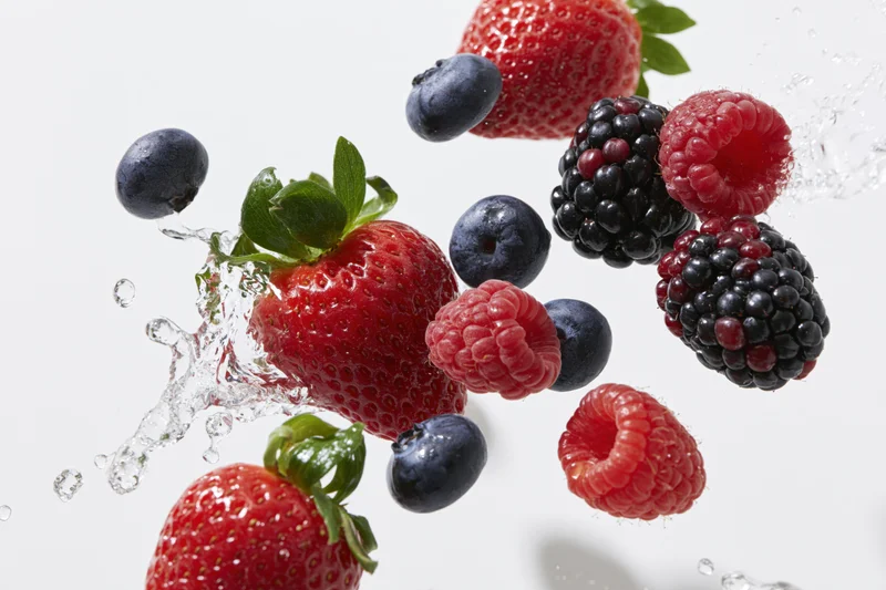Strawberries, blueberries, raspberries and blackberries suspended in mid-air with water splashing around them against a white background