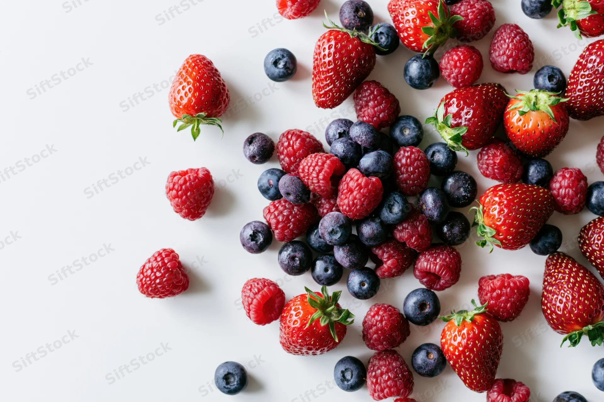 Mixed fresh strawberries raspberries and blueberries scattered on white surface with copyspace