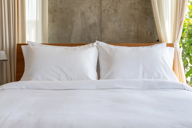 Neatly made hotel bed with white pillows and comforter against a concrete wall and window with curtains