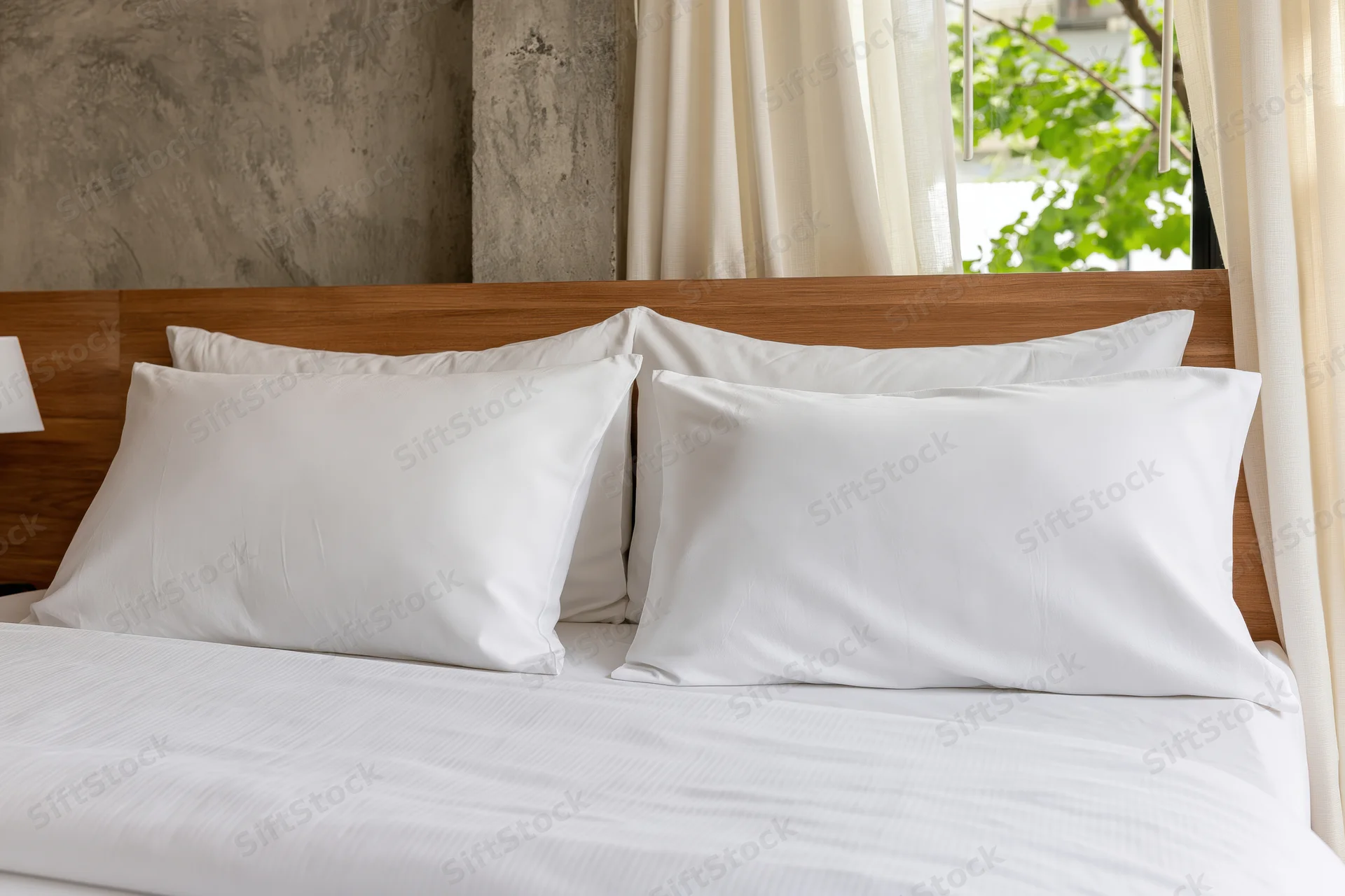 Neatly made hotel bed with four white pillows arranged against wooden headboard near window with curtains and greenery view