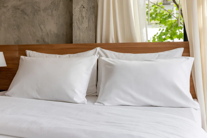 Neatly made hotel bed with four white pillows arranged against wooden headboard near window with curtains and greenery view