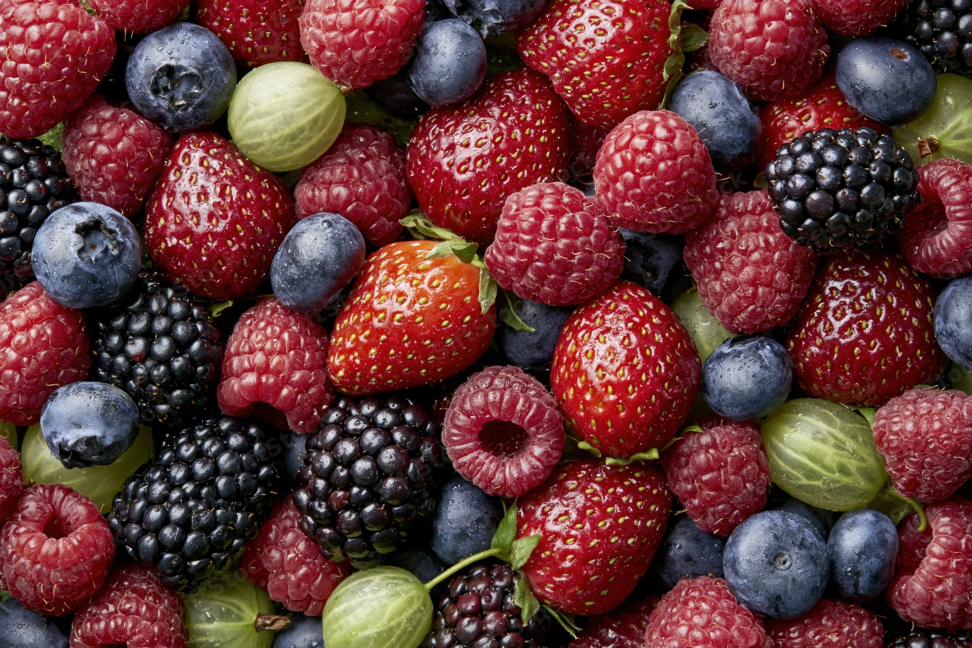 Overhead close-up of a dense pile of mixed fresh berries including strawberries, raspberries, blueberries, blackberries and gooseberries