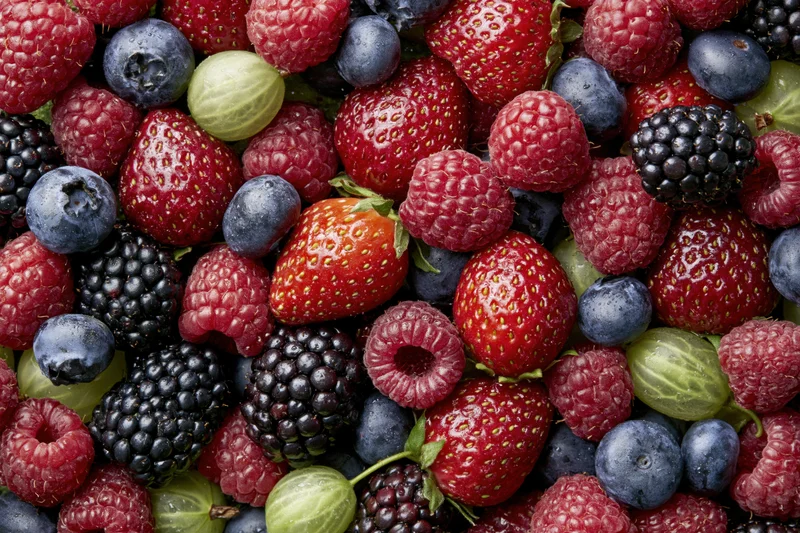 Overhead close-up of a dense pile of mixed fresh berries including strawberries, raspberries, blueberries, blackberries and gooseberries