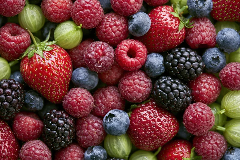 Overhead close-up of a dense pile of mixed fresh berries including strawberries, raspberries, blueberries, blackberries and gooseberries