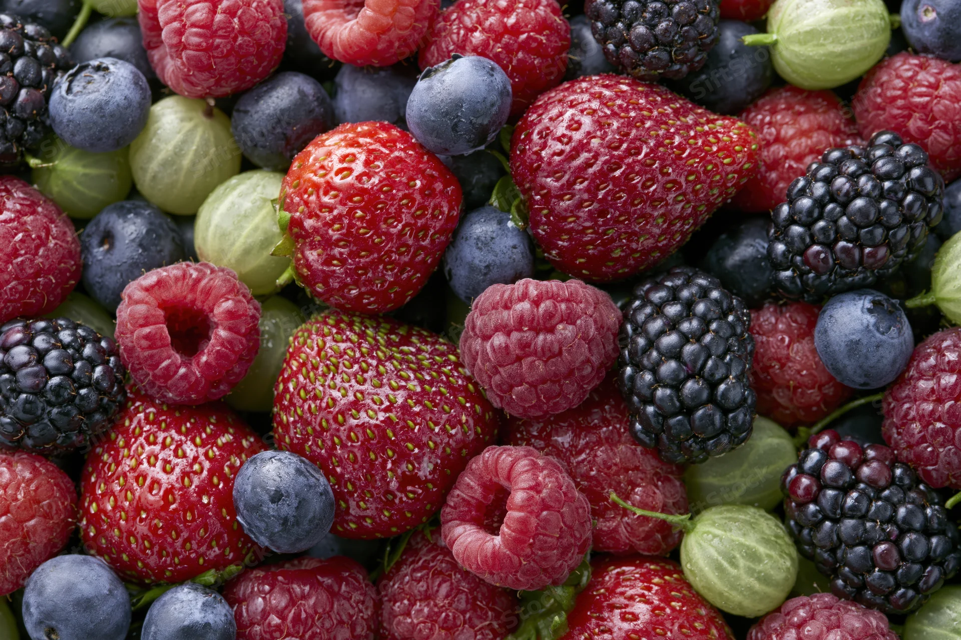 Overhead close-up of a dense pile of mixed fresh berries including strawberries, raspberries, blueberries, blackberries, and gooseberries