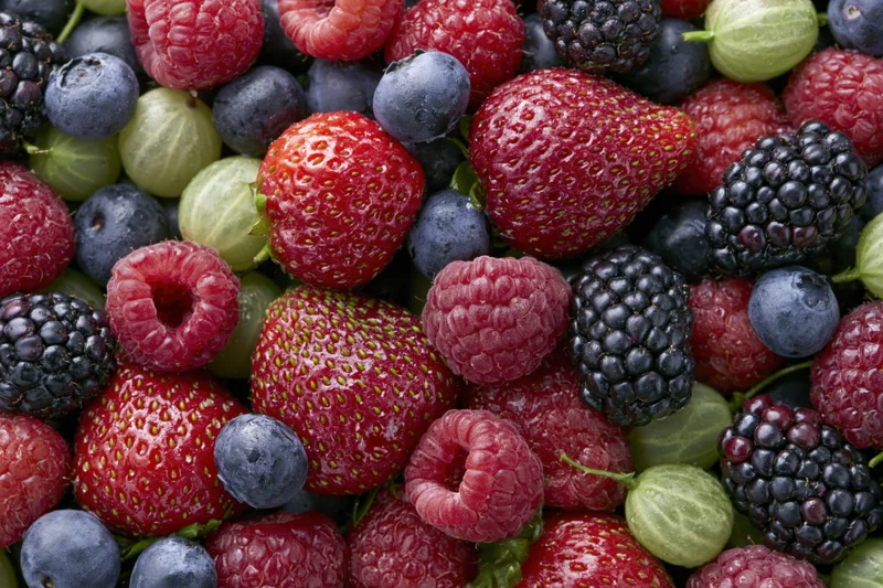 Overhead close-up of a dense pile of mixed fresh berries including strawberries, raspberries, blueberries, blackberries, and gooseberries