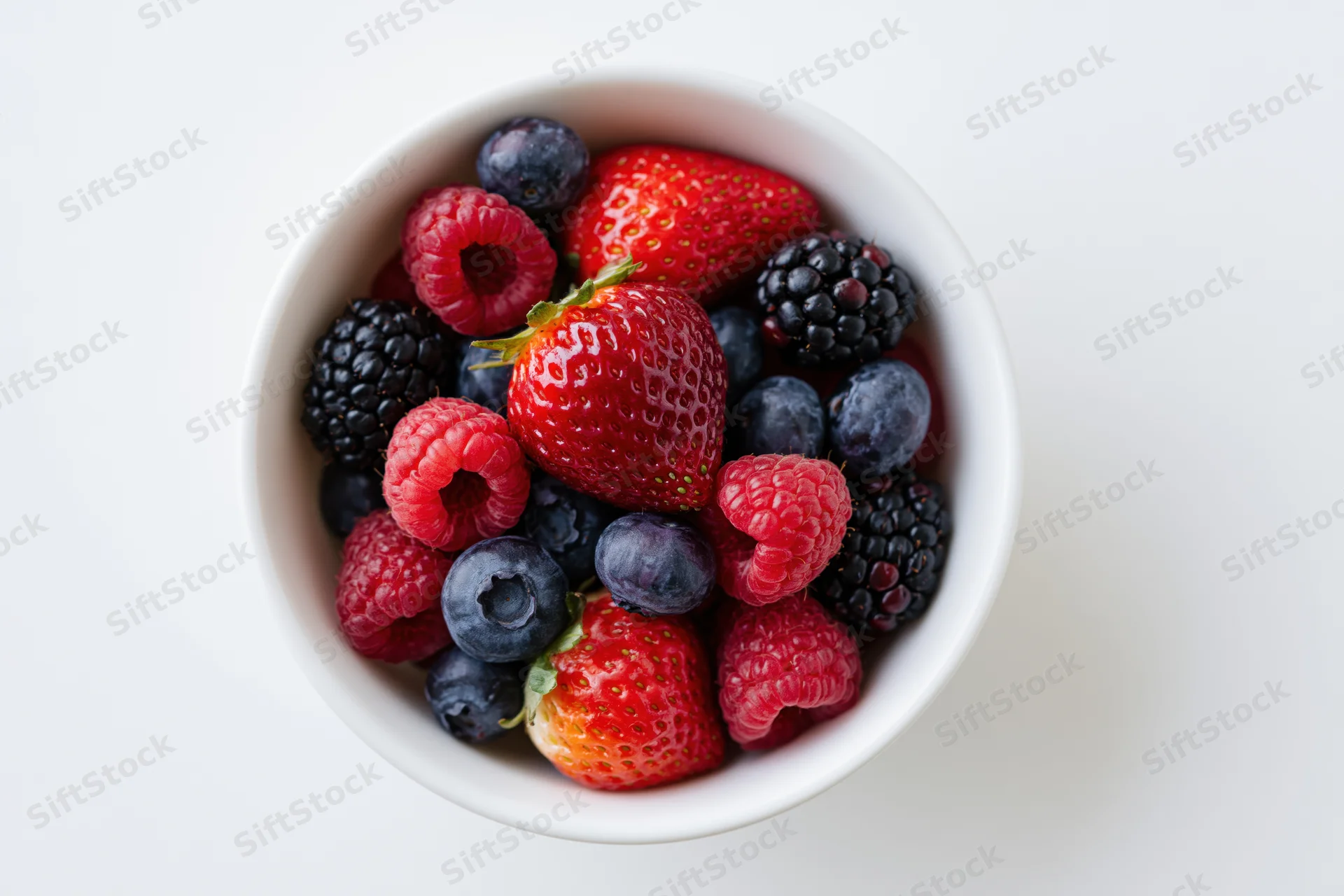 Overhead closeup of a white ceramic bowl filled with mixed berries on a white surface