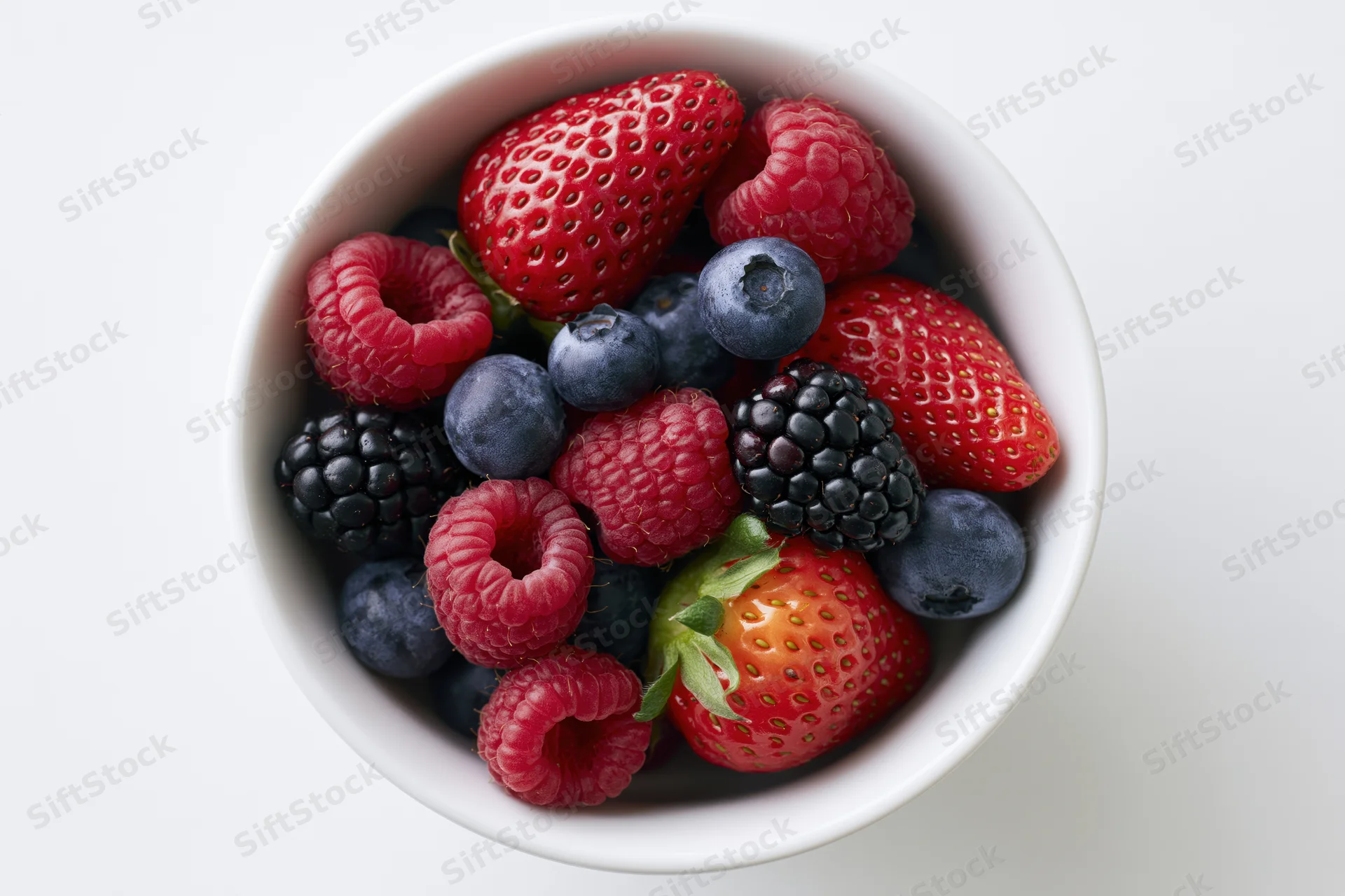 Overhead close-up of a white ceramic bowl filled with mixed berries including strawberries, raspberries, blueberries, and blackberries on a plain white surface