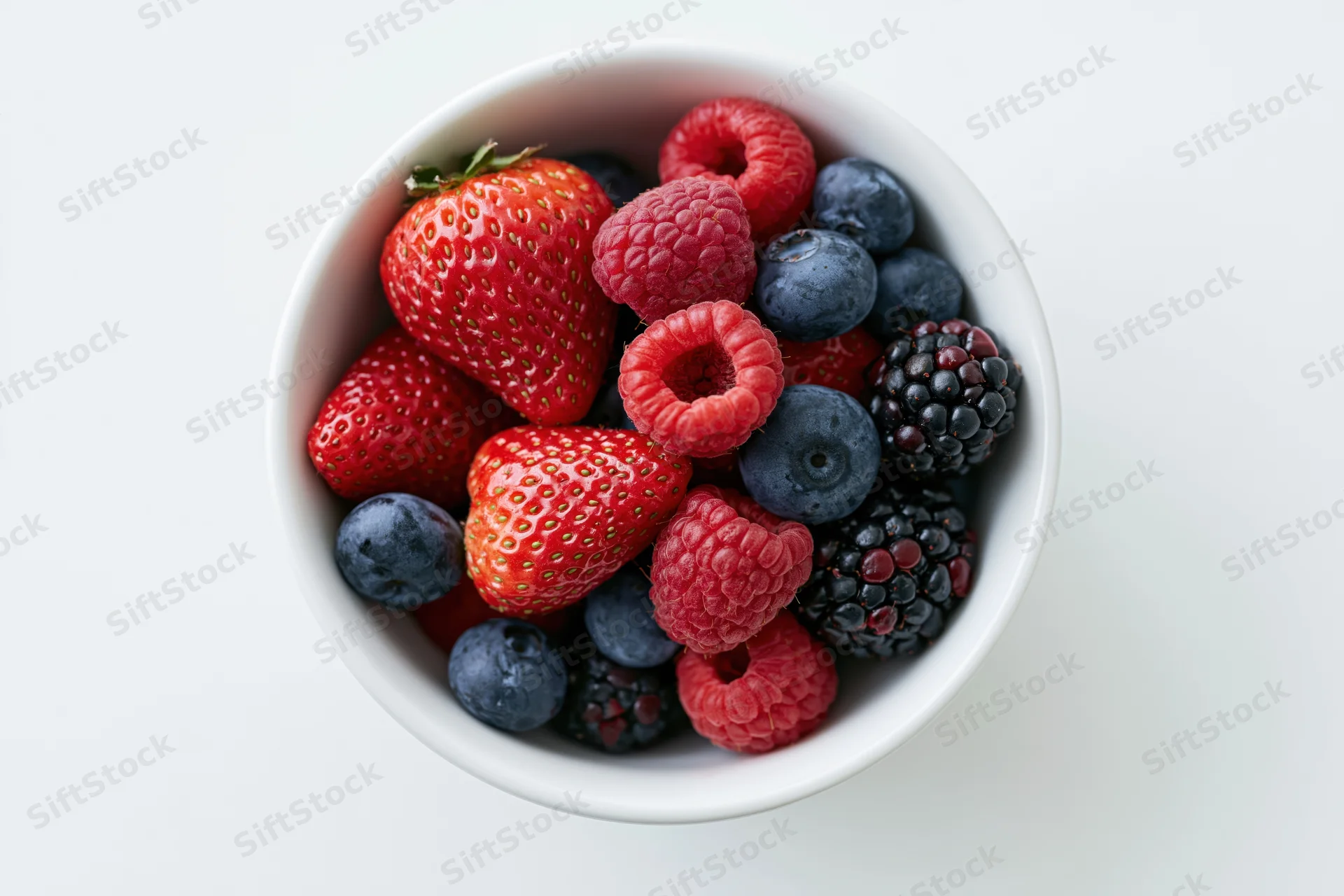Overhead closeup of a white ceramic bowl filled with strawberries, raspberries, blueberries and blackberries on a white surface