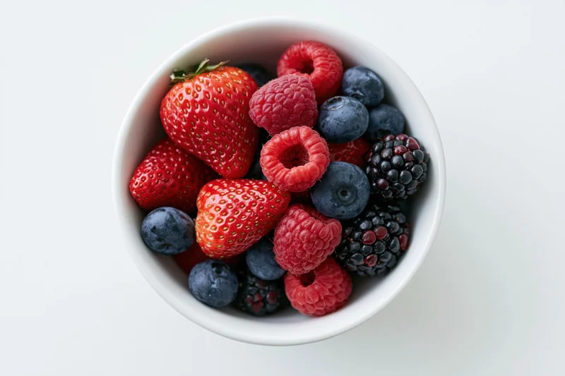 Overhead closeup of a white ceramic bowl filled with strawberries, raspberries, blueberries and blackberries on a white surface