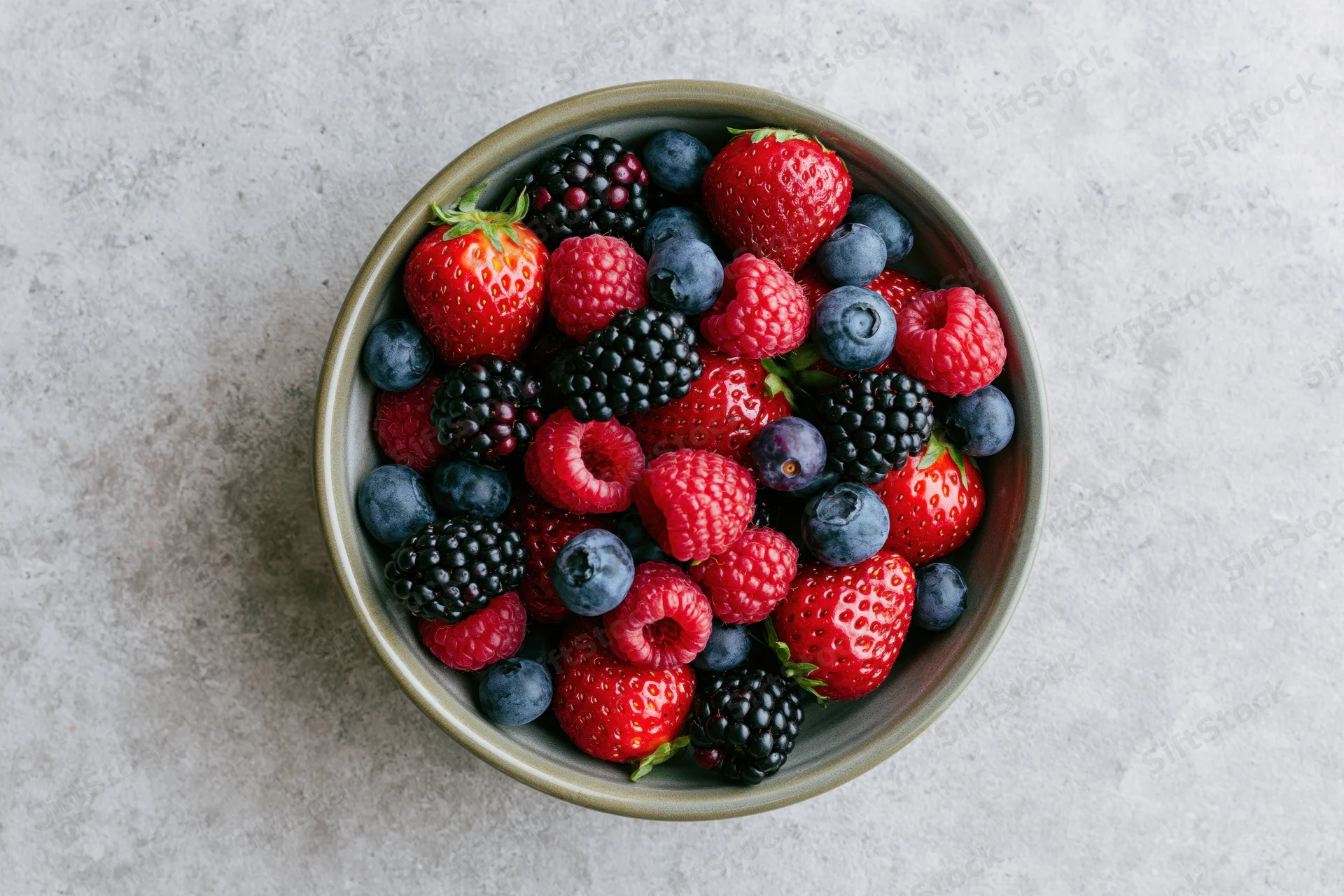 Overhead view of a ceramic bowl filled with mixed fresh berries on a gray stone surface