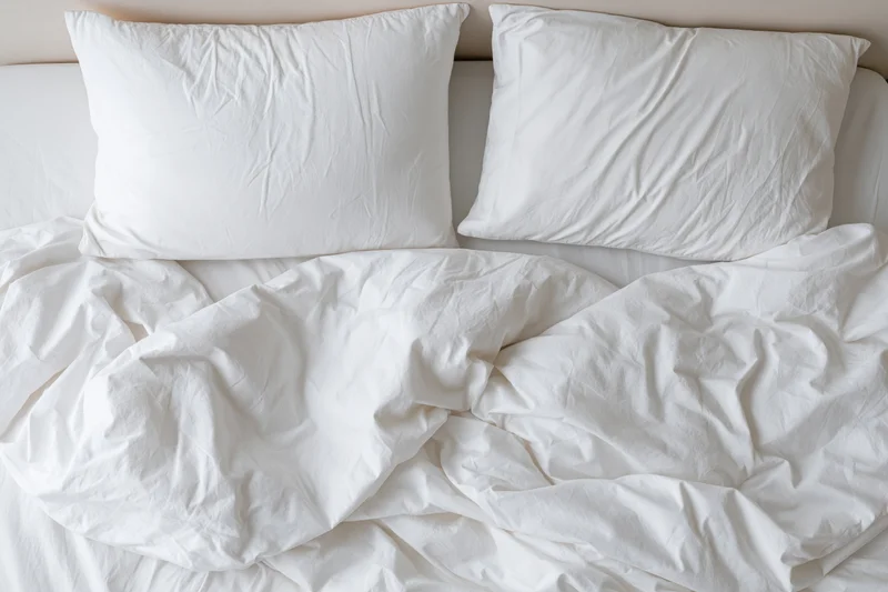 Overhead view of unmade bed with two white pillows and crumpled white sheets and comforter