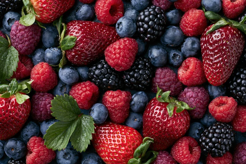 Overhead flat-lay of mixed fresh berries including strawberries, blueberries, raspberries, and blackberries with green leaves and water droplets