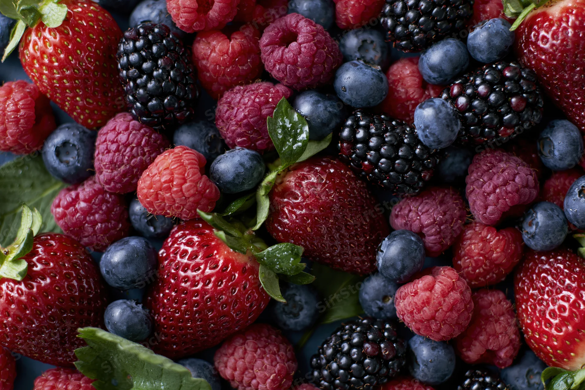 Overhead flat-lay of mixed fresh berries including strawberries, blueberries, raspberries and blackberries on a dark surface