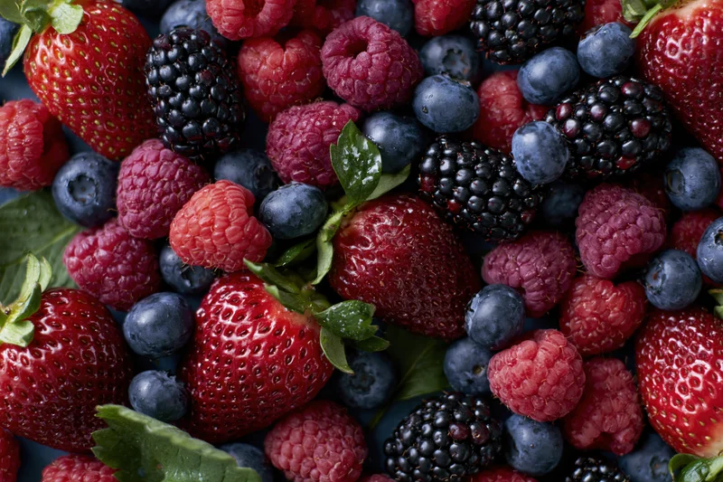 Overhead flat-lay of mixed fresh berries including strawberries, blueberries, raspberries and blackberries on a dark surface