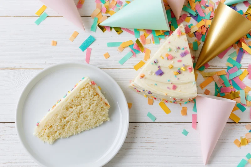 Slice of birthday cake with frosting and sprinkles on white plate beside party hats and confetti on wooden table
