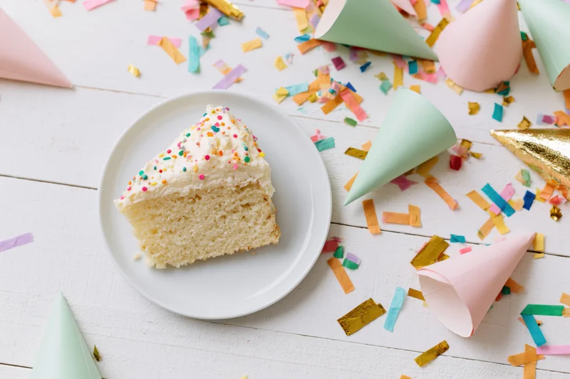 Slice of birthday cake with sprinkles on white plate surrounded by party hats and confetti on wooden surface